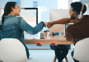 Rearview shot of two young designers giving each other a fist bump in an office, on display is Crestron desk scheduling device