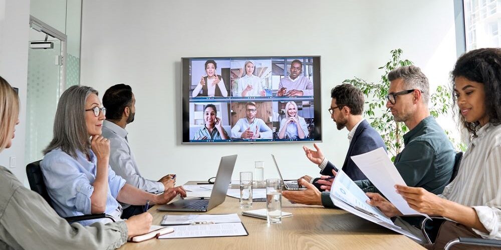 Workers in a conference room with a screen showing remote participants on Zoom.