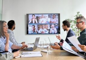 Workers in a conference room with a screen showing remote participants on Zoom.